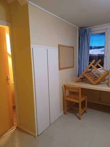 Petite chambre avec une armoire blanche, un bureau et une chaise en bois, des rideaux bleus et une fenêtre donnant sur l'extérieur.