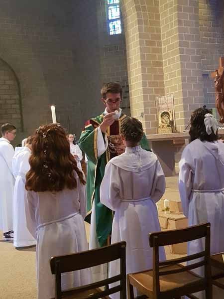 Un prêtre en vêtements liturgiques verts s'adresse à des enfants en robes blanches pendant un office religieux.