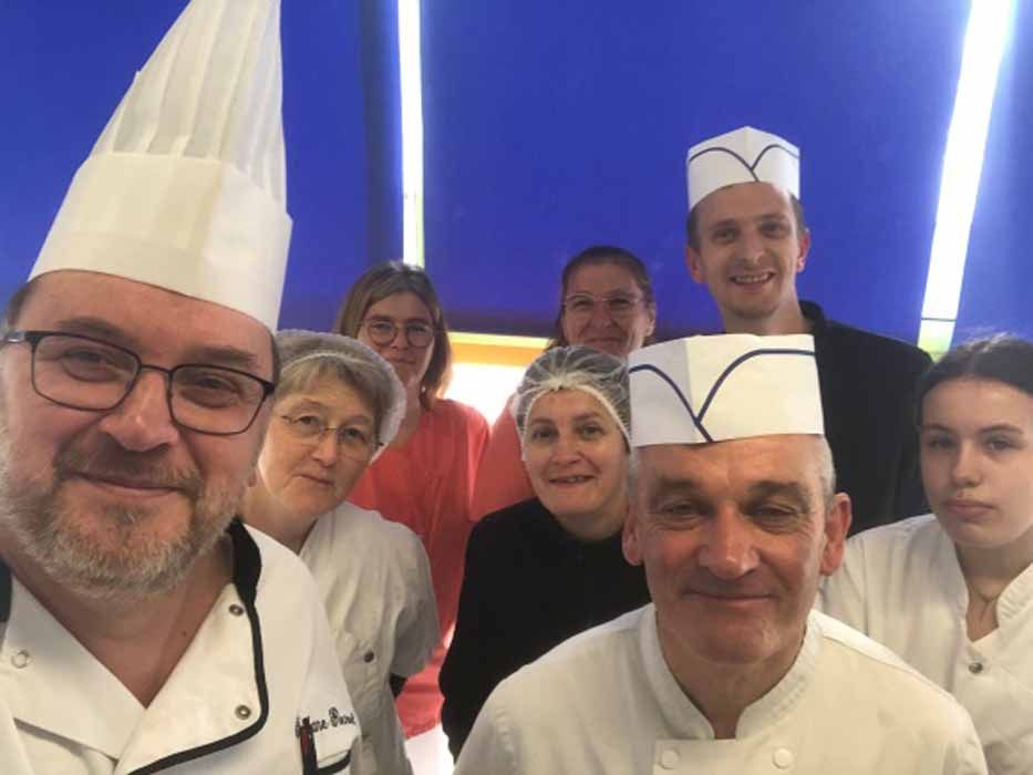 Un groupe de chefs en toques blanches prend un selfie dans une cuisine, souriant à l'objectif.