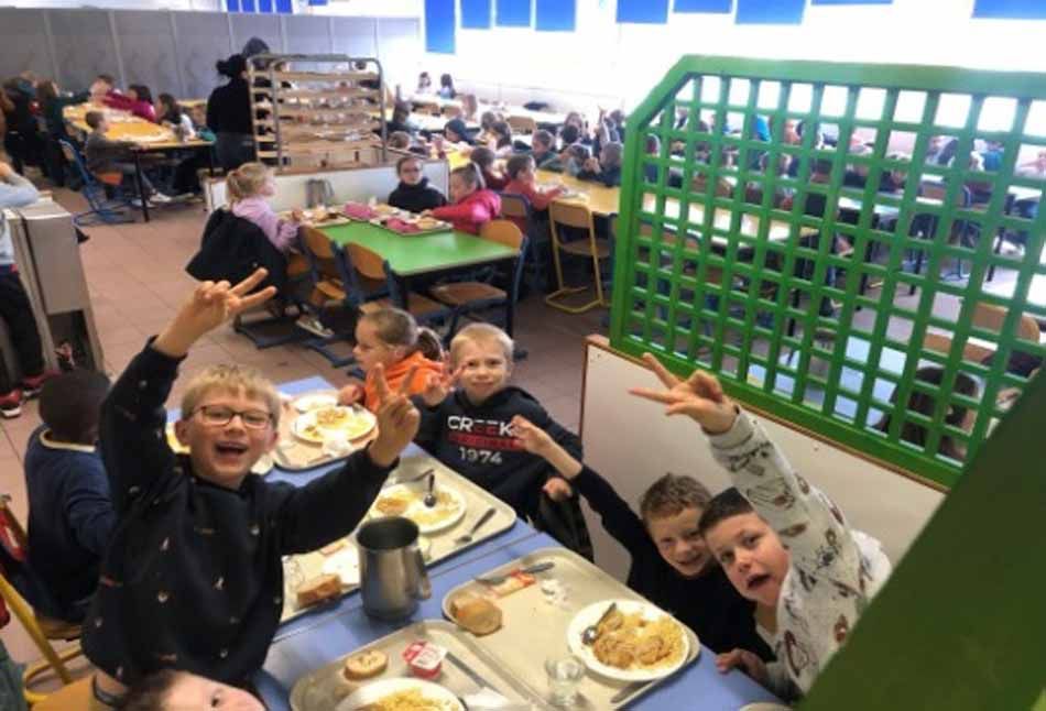 Des enfants assis à une table de cafétéria, souriant et faisant signe à la caméra