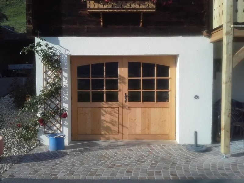 Porte de garage double en bois dans un mur blanc, encadrée de plantes en pot sur une allée pavée.