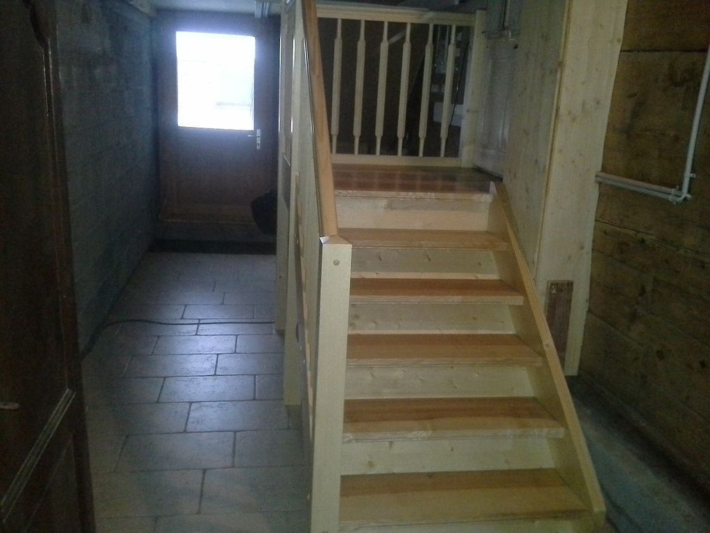 Escalier intérieur étroit avec marches beiges, une rampe et un couloir carrelé menant à une porte lumineuse.