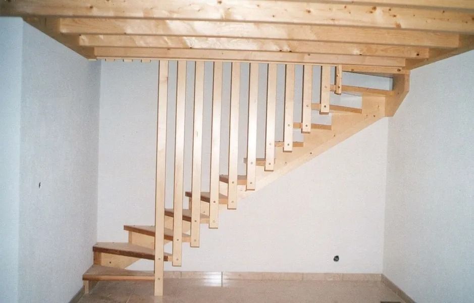 Escalier en bois à lattes verticales sous poutres apparentes au plafond dans une pièce blanche