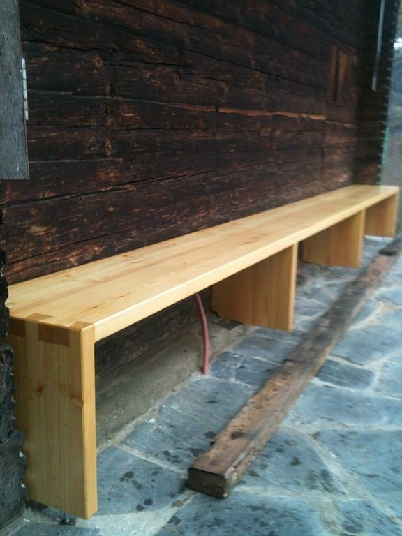 Long banc en bois adossé à un mur sombre sur un sol en pierre, avec une planche mal fixée devant.