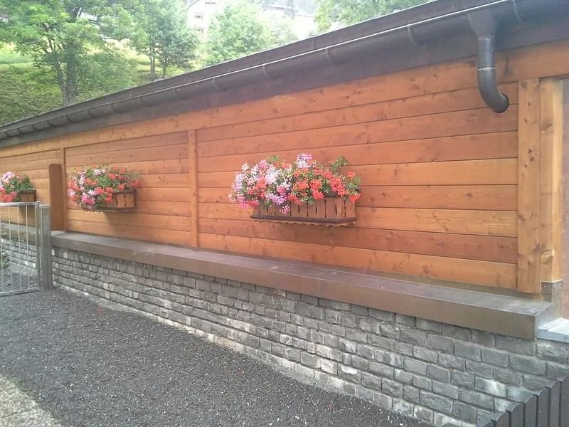 Clôture en bois ornée de paniers de fleurs au-dessus d'un muret en pierre, le long d'un chemin de gravier.