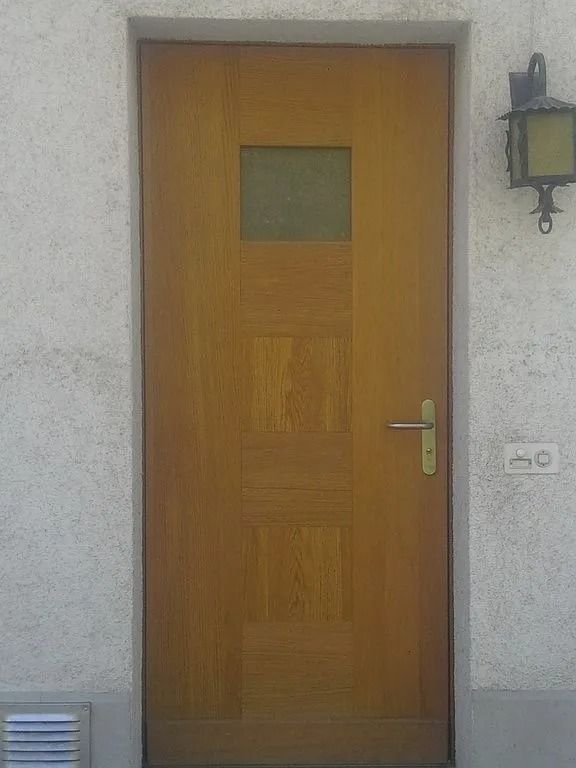 Porte d'entrée en bois avec une petite fenêtre, encastrée dans un mur extérieur gris clair avec une applique murale.