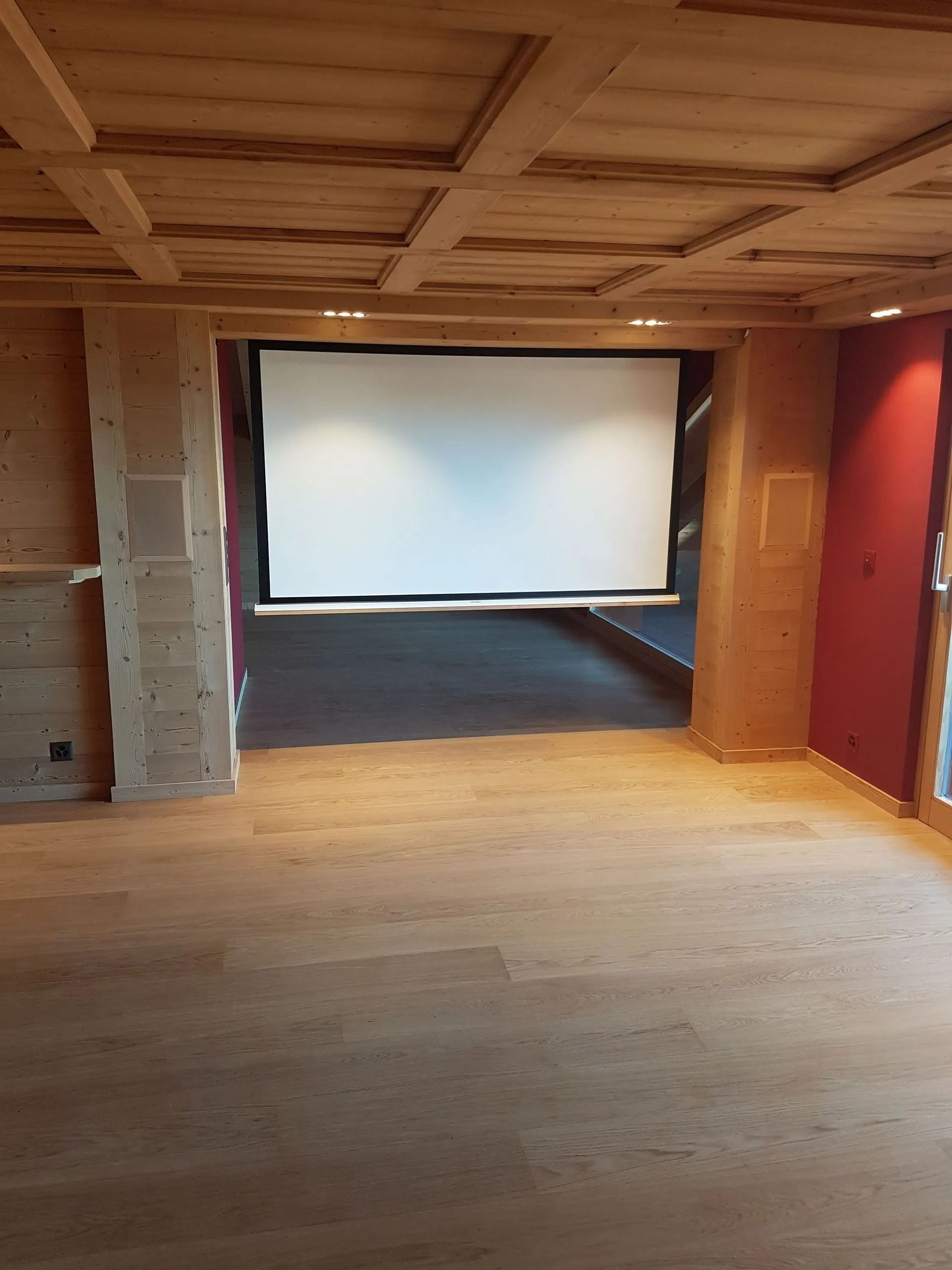 Pièce vide et inachevée avec parquet, poutres apparentes et une grande fenêtre donnant sur un extérieur lumineux.