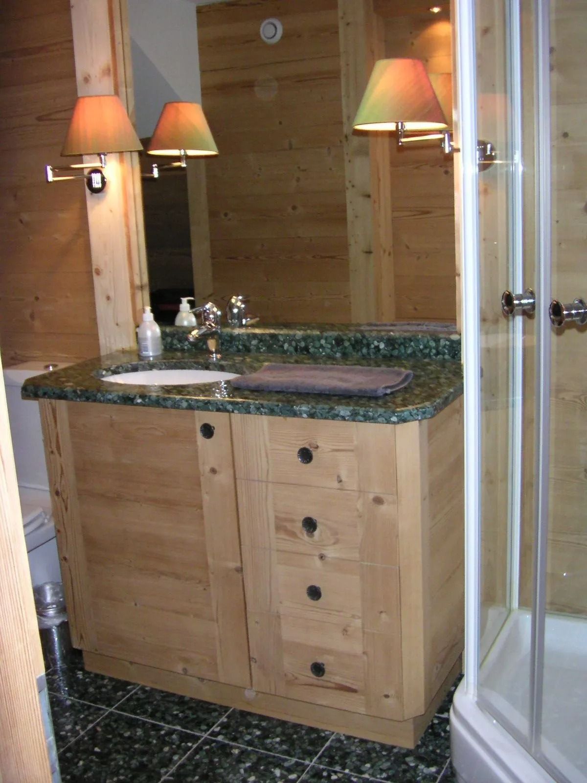 Meuble-lavabo en bois avec plateau en marbre vert, miroir, lavabo et cabine de douche attenante.