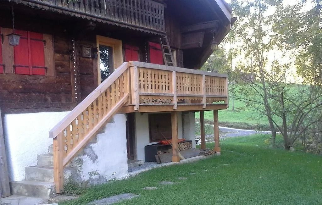 Balcon et escalier en bois attenants à une maison rustique, à côté d'une cour gazonnée et d'arbres.