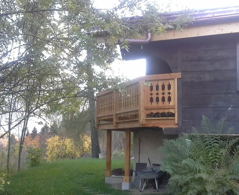 Balcon en bois sur une maison bordée de conifères et d'une cour gazonnée.