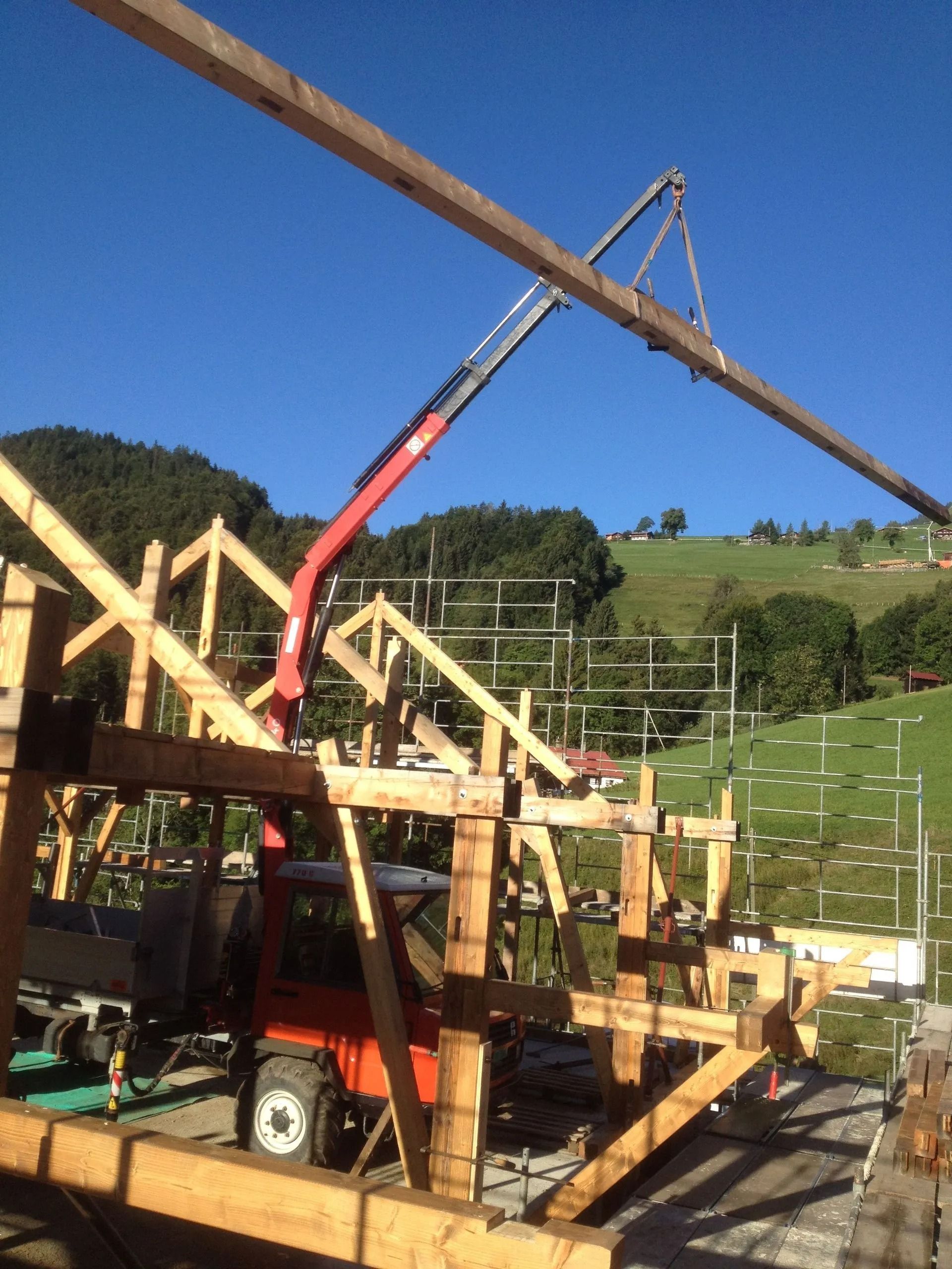 Une grue rouge soulève des poutres en bois pour la charpente d'une maison sur un chantier de construction, sous un ciel bleu.