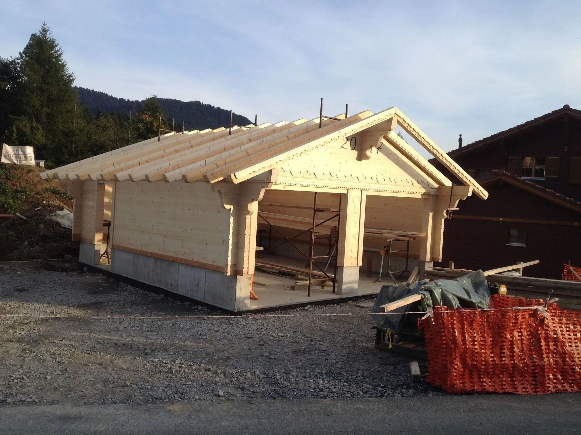 Maison à ossature bois partiellement construite sur un terrain gravillonné, avec une clôture de sécurité orange et des montagnes en arrière-plan.