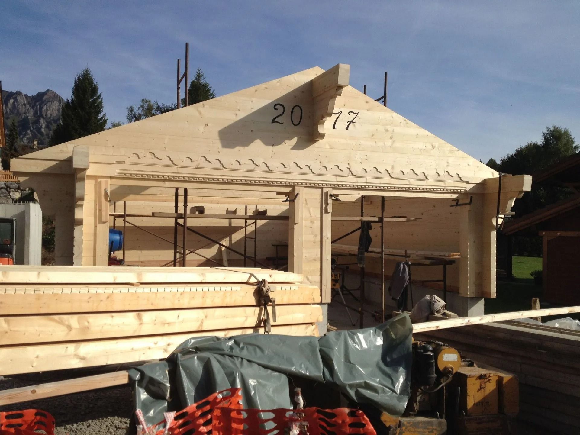 Maison en construction avec charpente apparente, piles de bois et bâches dans un quartier résidentiel