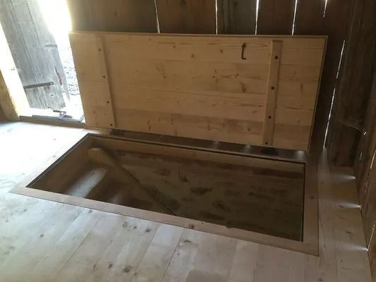 Trappe en bois s'ouvrant dans un plancher rustique, révélant un vide sanitaire ou un sous-sol sombre en dessous.