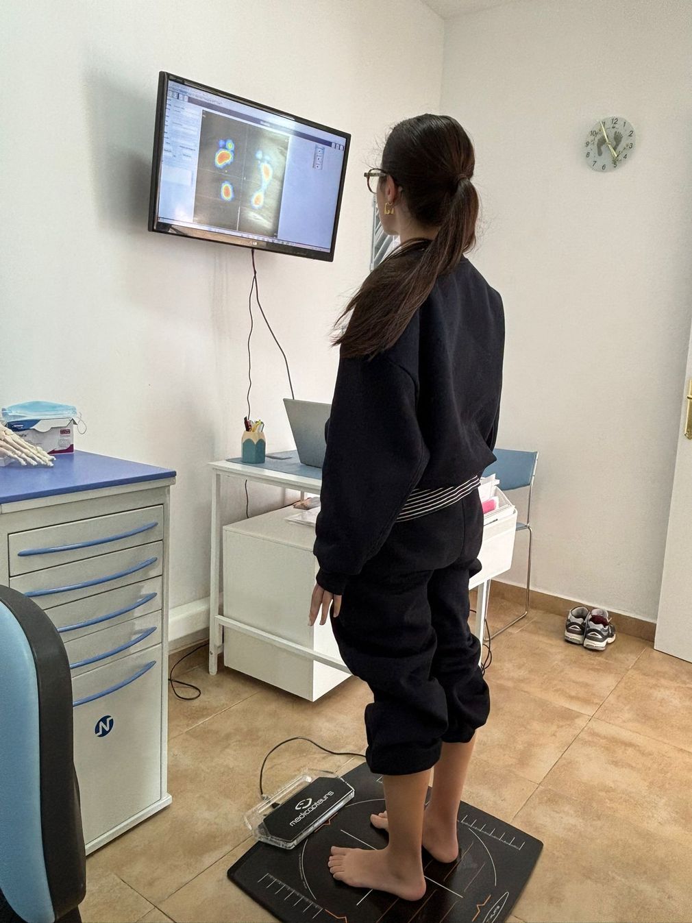 Persona de pie sobre una plataforma de equilibrio en una clínica, frente a una pantalla montada en la pared durante una evaluación médica.