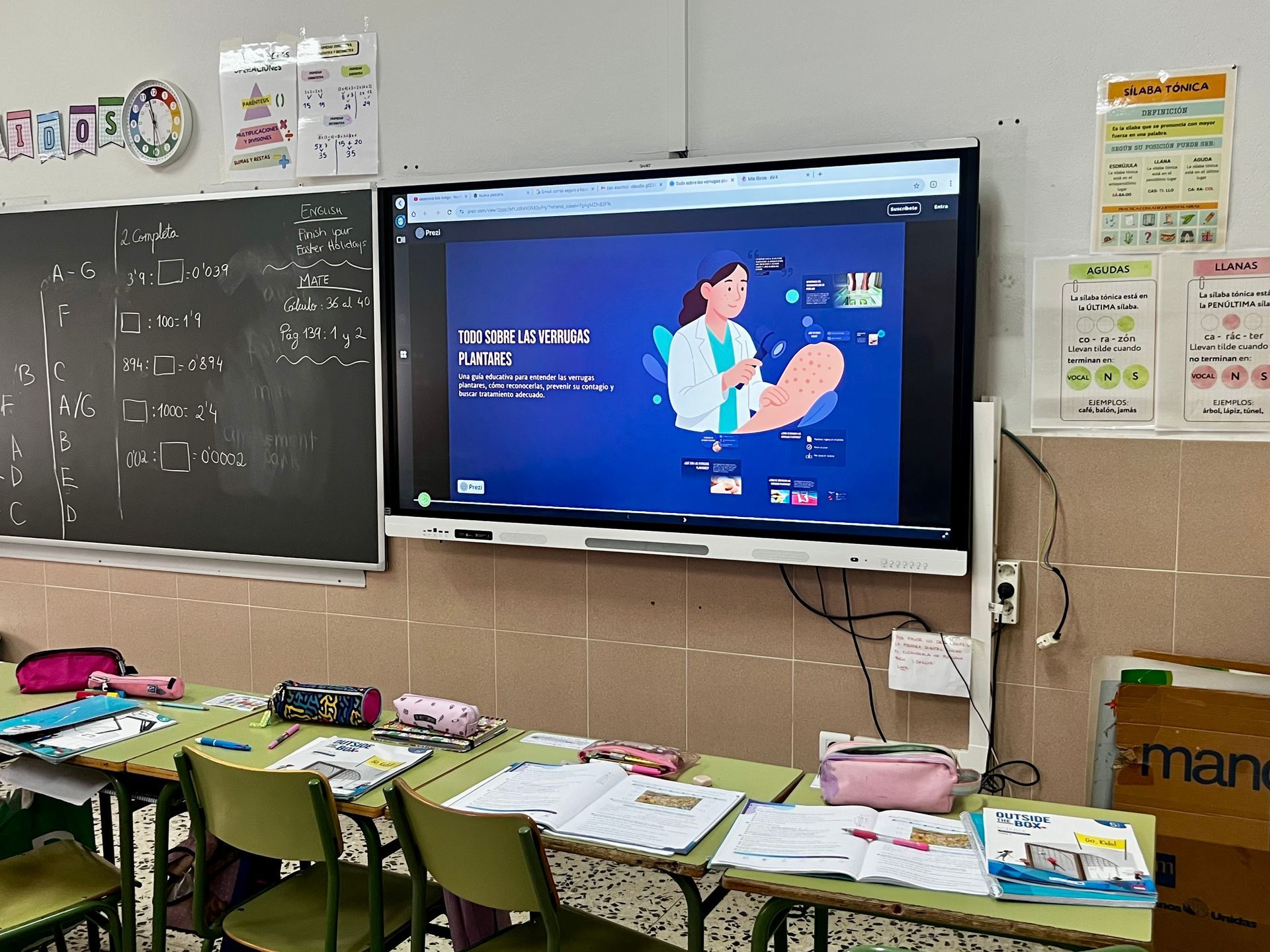 Aula con pizarra, televisor montado en la pared que muestra dibujos animados y pupitres para estudiantes con libros y material escolar.