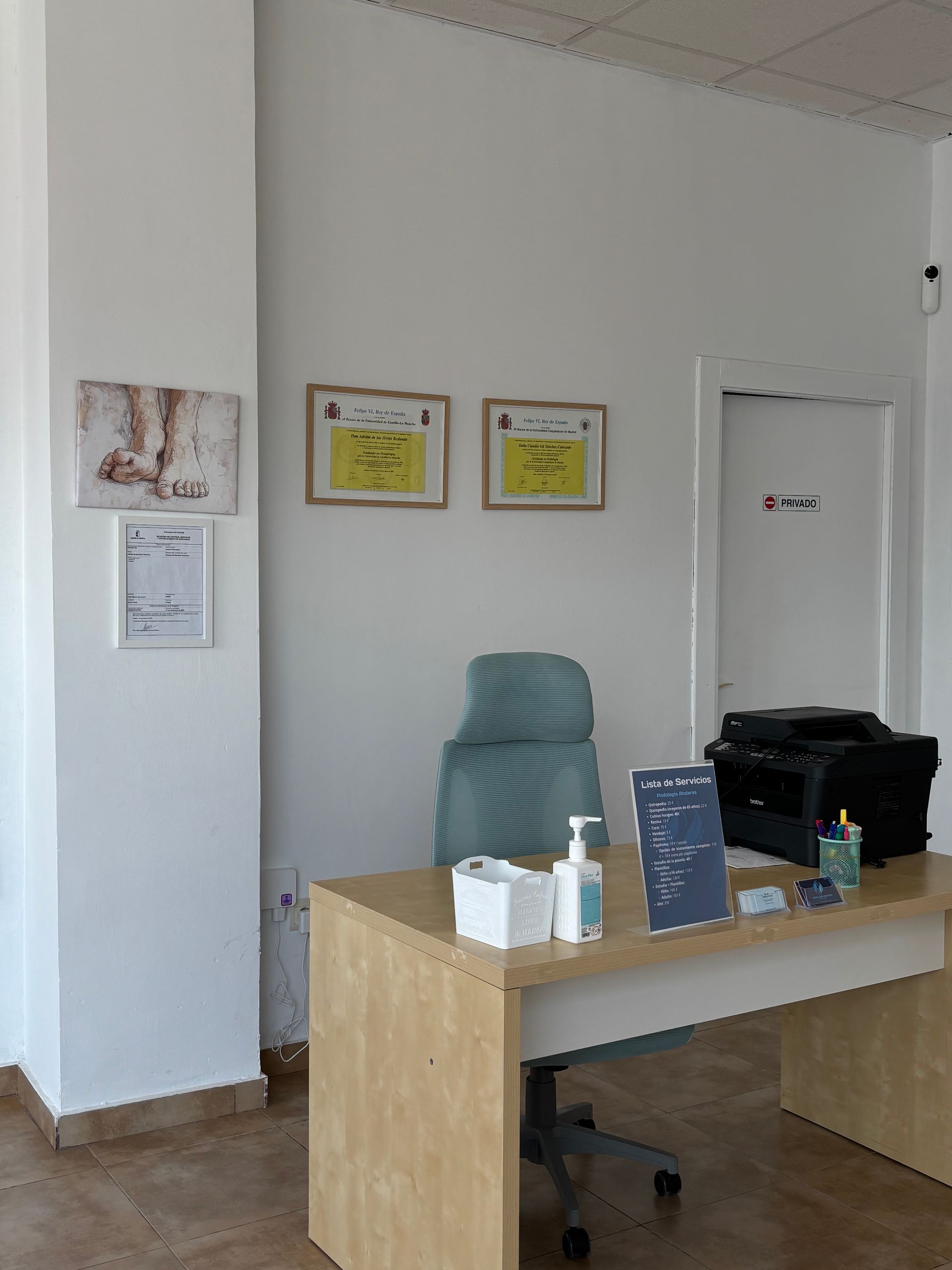 Mostrador de recepción de oficina con sillas apiladas, avisos enmarcados y una impresora contra una pared blanca.