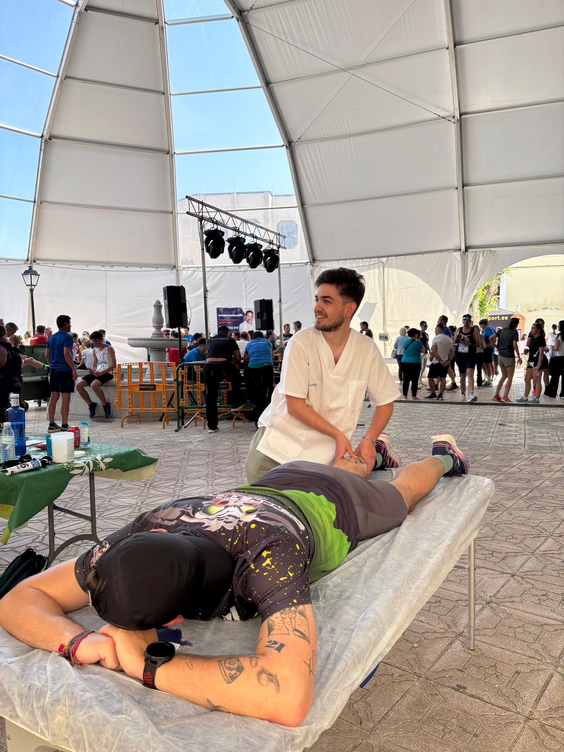 Persona recibiendo un masaje de espalda sobre una colchoneta en un gran espacio para eventos cubierto con una carpa.