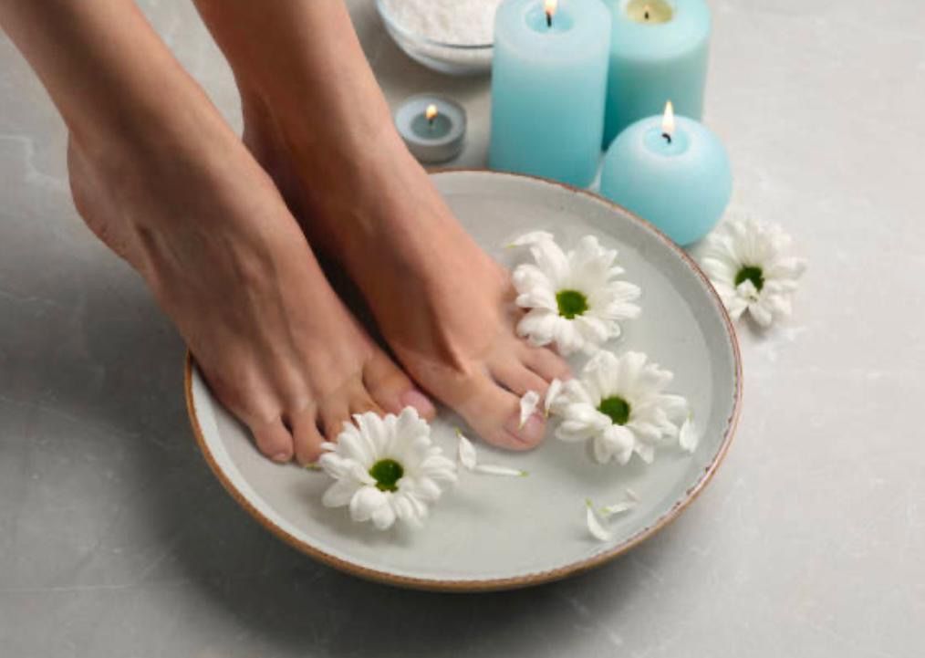 Füße, die in einer Schüssel mit Wasser, weißen Blüten, Kerzen und Salz auf grauem Untergrund baden.