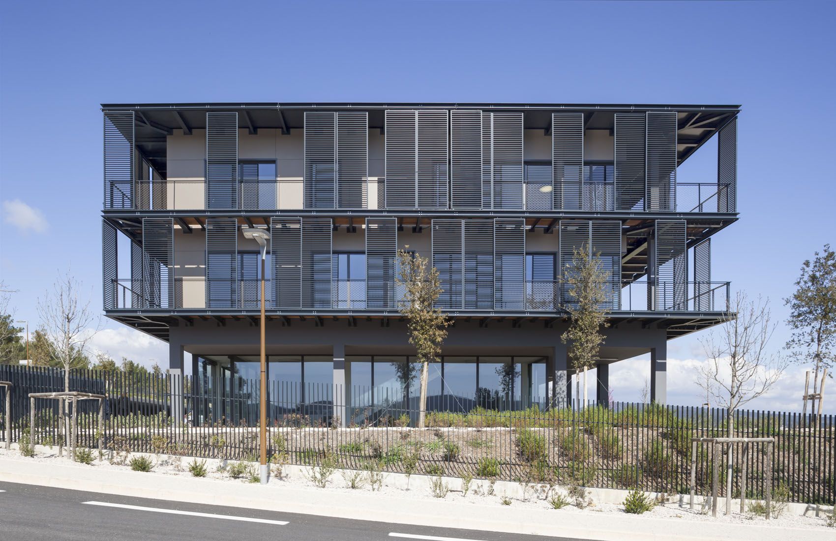 Immeuble d'appartements moderne de trois étages avec balcons et façade métallique, entouré d'aménagements paysagers sous un ciel bleu azur.