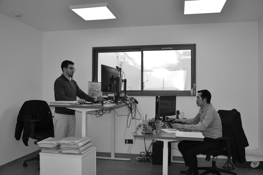 Deux personnes travaillent à des bureaux dans un bureau lumineux avec de grandes fenêtres et des écrans d'ordinateur.