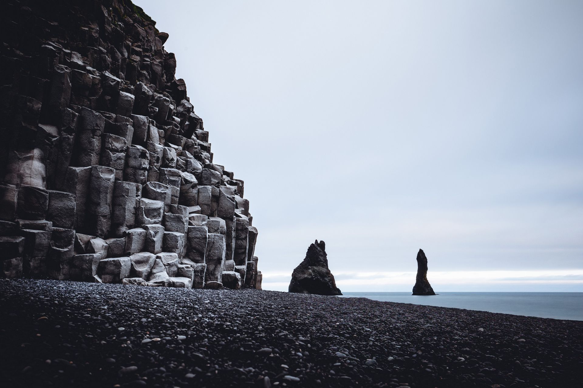 Columnas de basalto negro junto a una playa de arena negra, dos farallones en el océano, cielo gris.