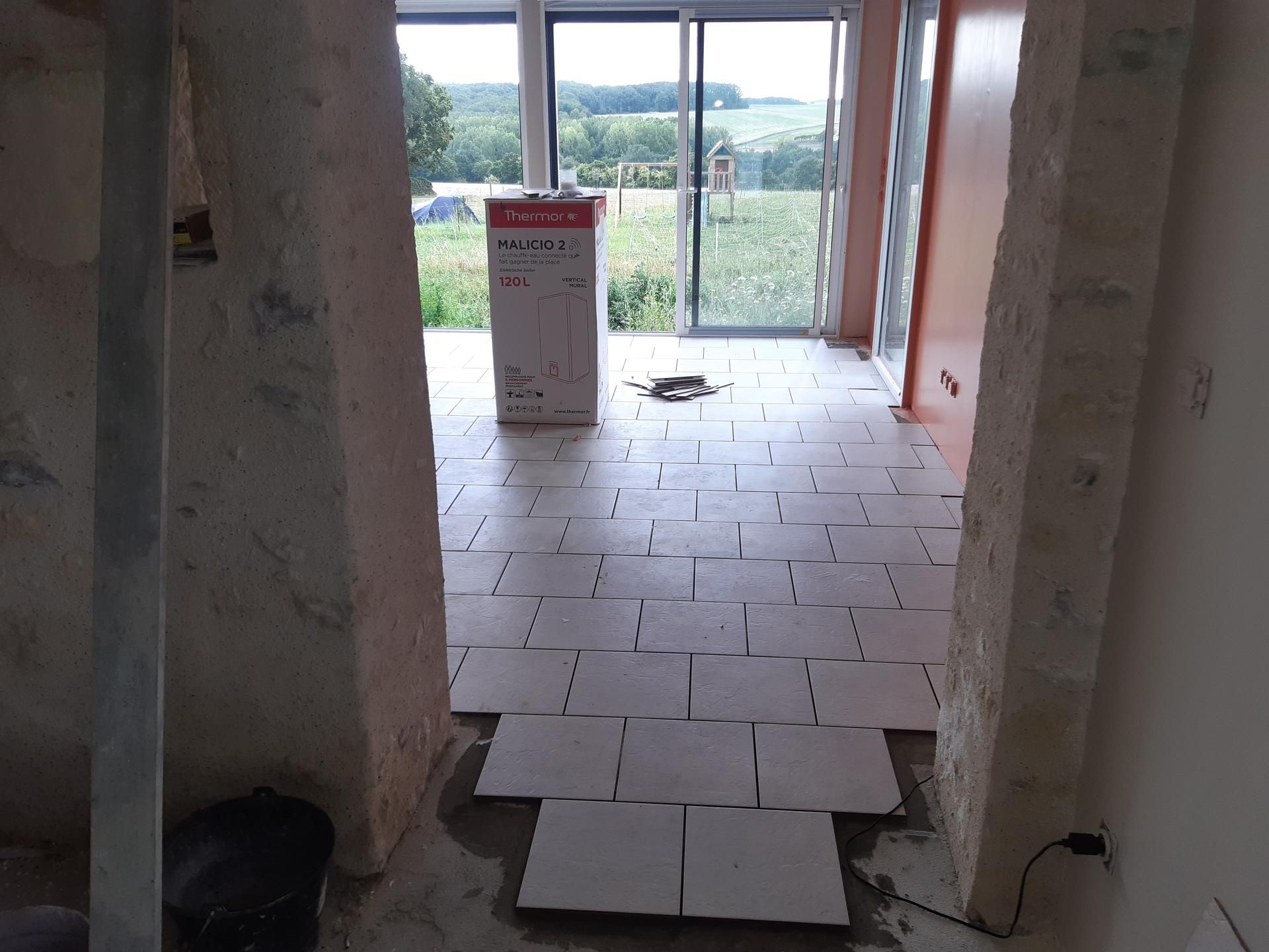 Vue intérieure d'une pièce avec sol partiellement carrelé ; en arrière-plan, porte coulissante en verre donnant sur l'extérieur.