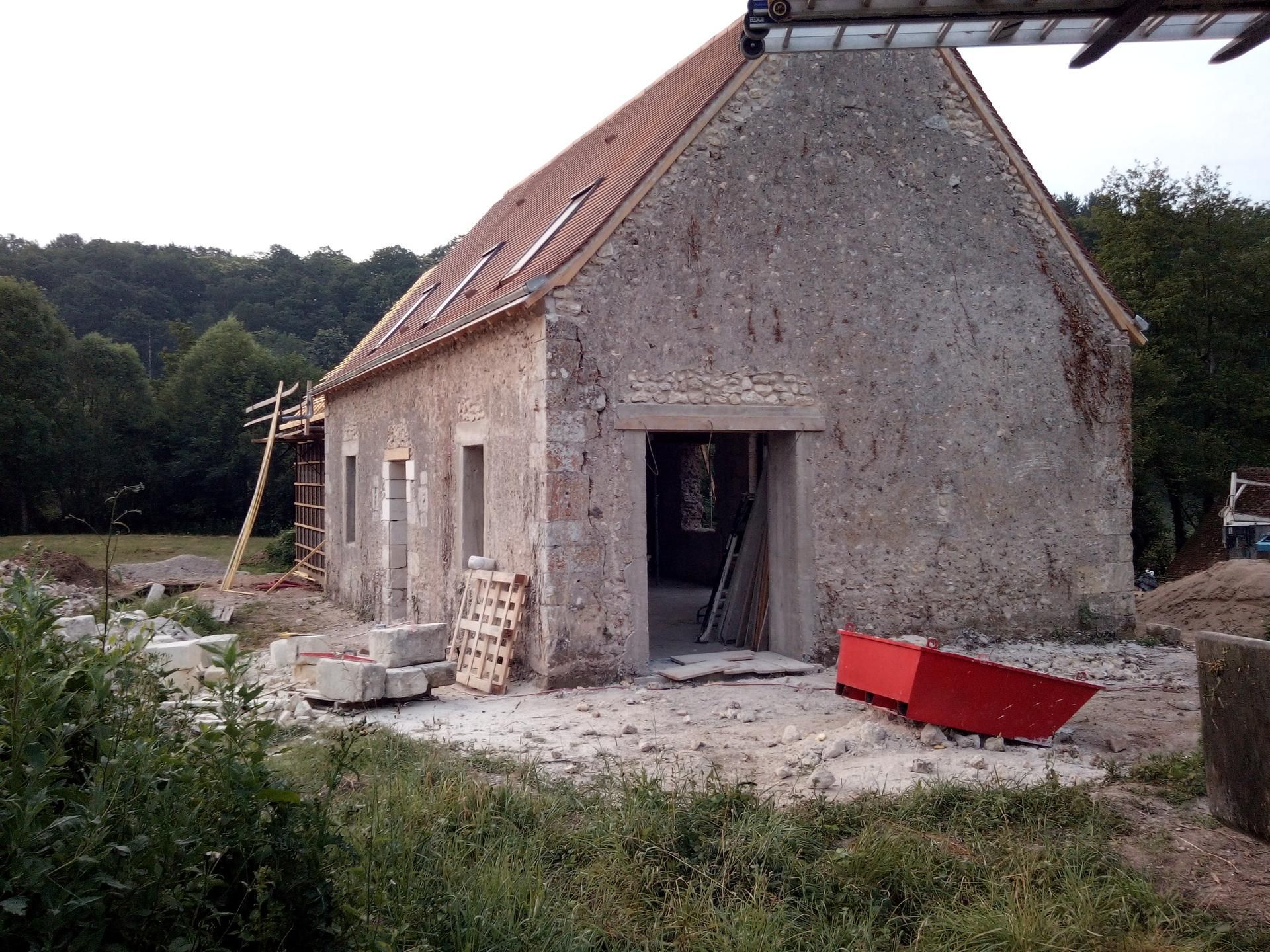 Bâtiment en pierre en rénovation ; toit rouge, porte ouverte, matériaux de construction à proximité.