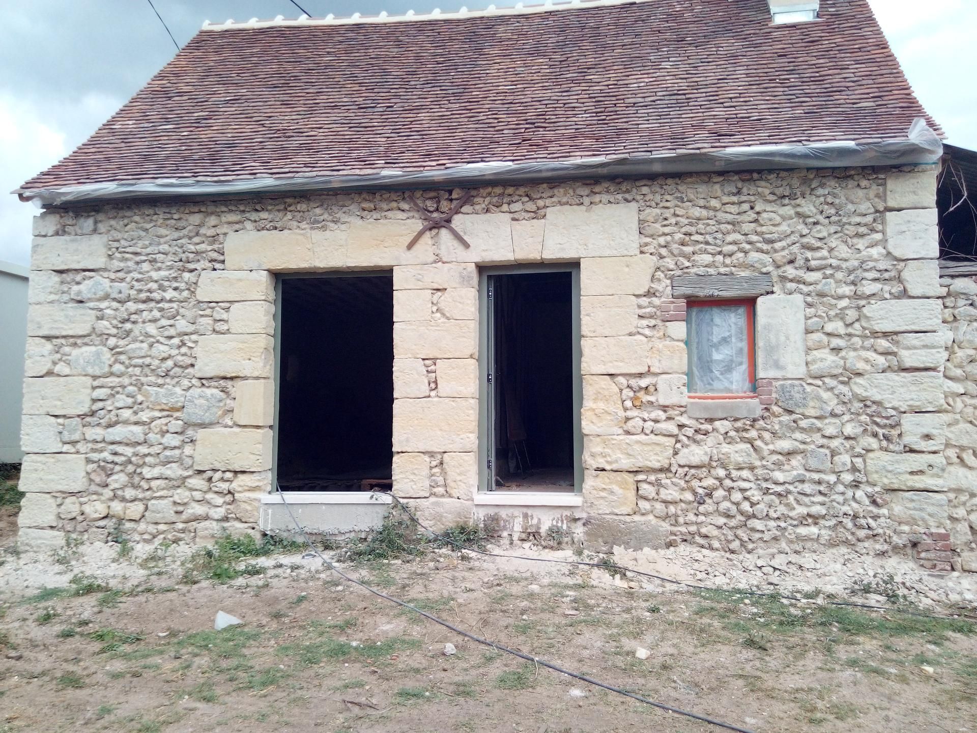 Maison en pierre avec portes ouvertes, petite fenêtre et toit de tuiles rouges.