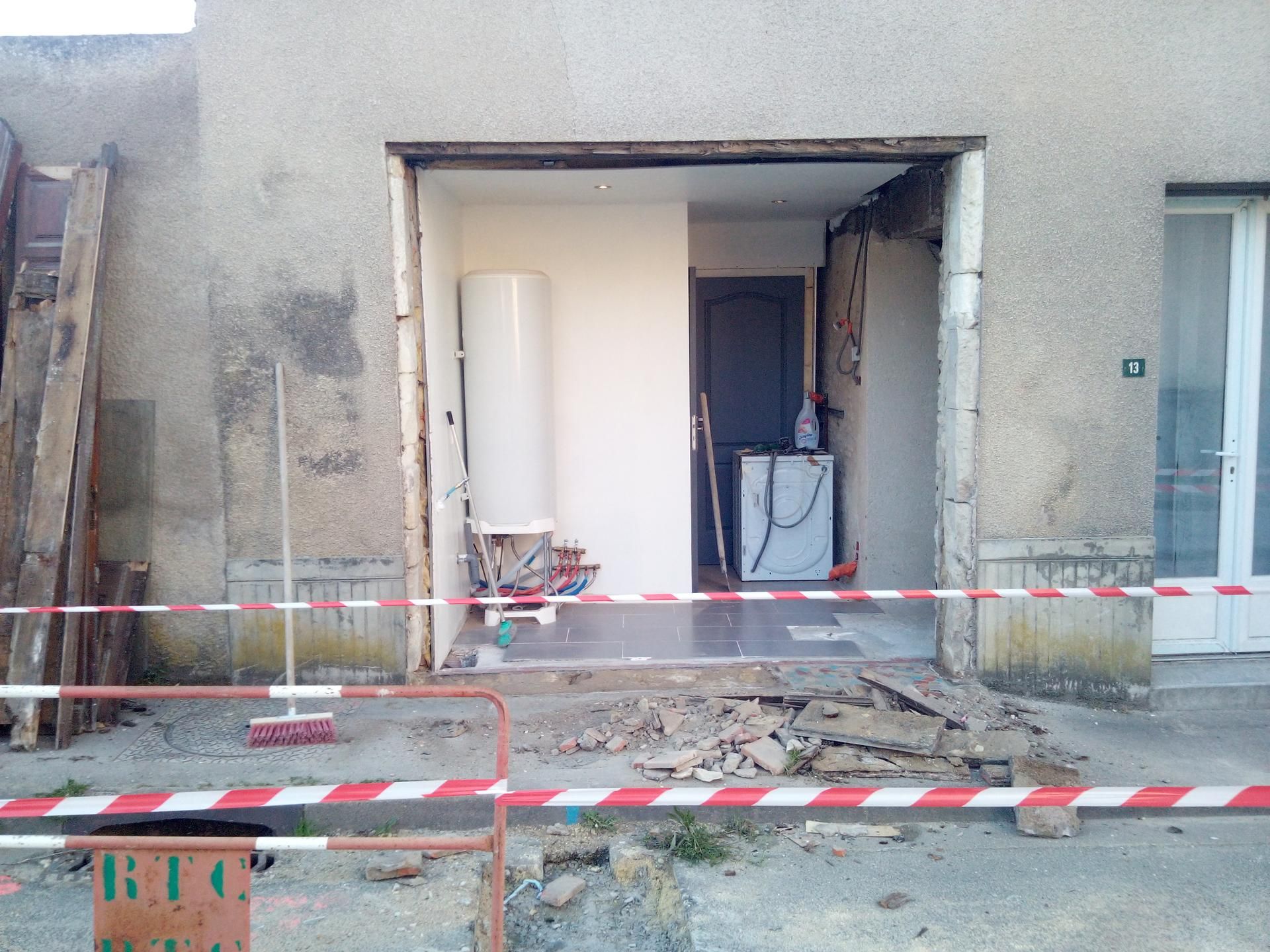 Vue extérieure d'un bâtiment en construction, porte ouverte, chauffe-eau visible, débris au sol, ruban de signalisation rouge et blanc.