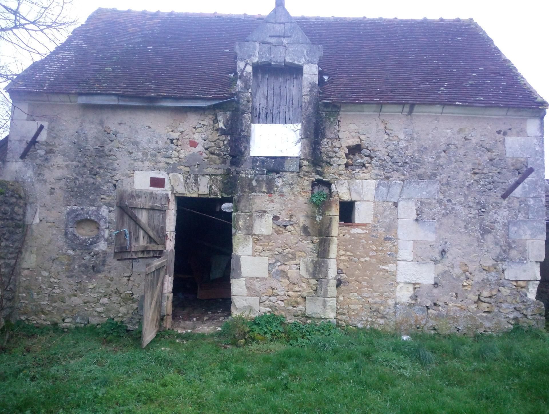Ancien bâtiment en pierre avec un toit de tuiles rouges, une porte ouverte et une porte en bois ; situé dans un espace herbeux.