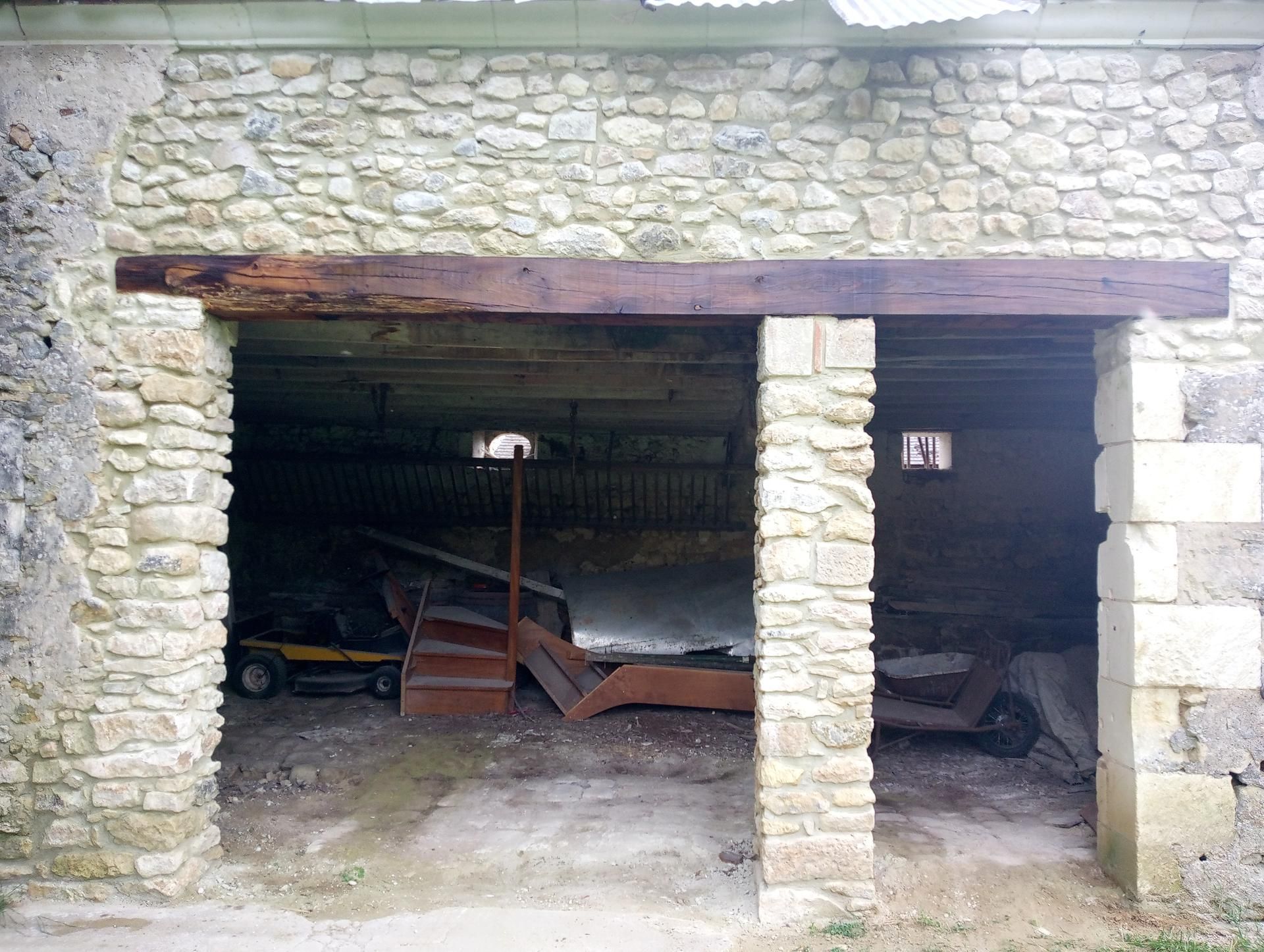 Bâtiment en pierre avec deux portes ouvertes, une poutre en bois et de vieux équipements à l'intérieur.