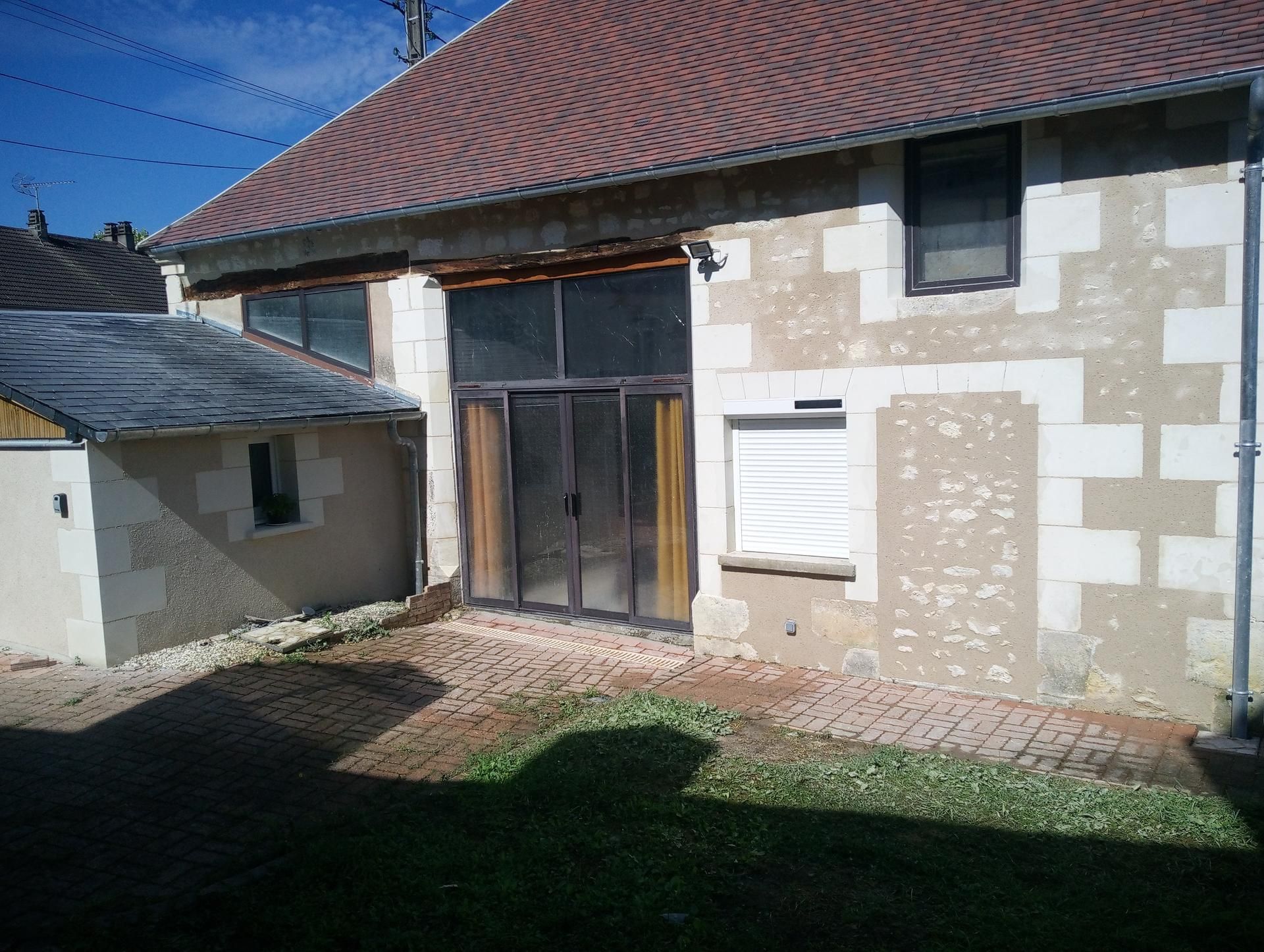 Bâtiment en pierre avec toit de tuiles rouges, porte vitrée et petite dépendance sous un ciel bleu.
