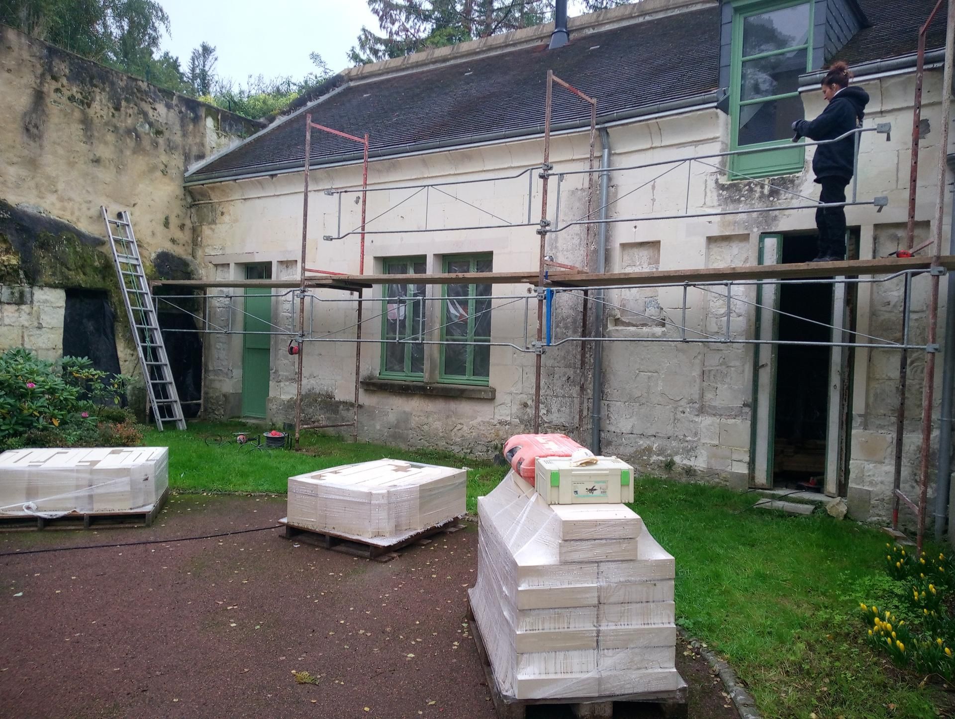 Extérieur d'un bâtiment en rénovation avec des échafaudages. Une personne travaille sur une fenêtre. Des palettes de blocs de pierre sont posées au sol.