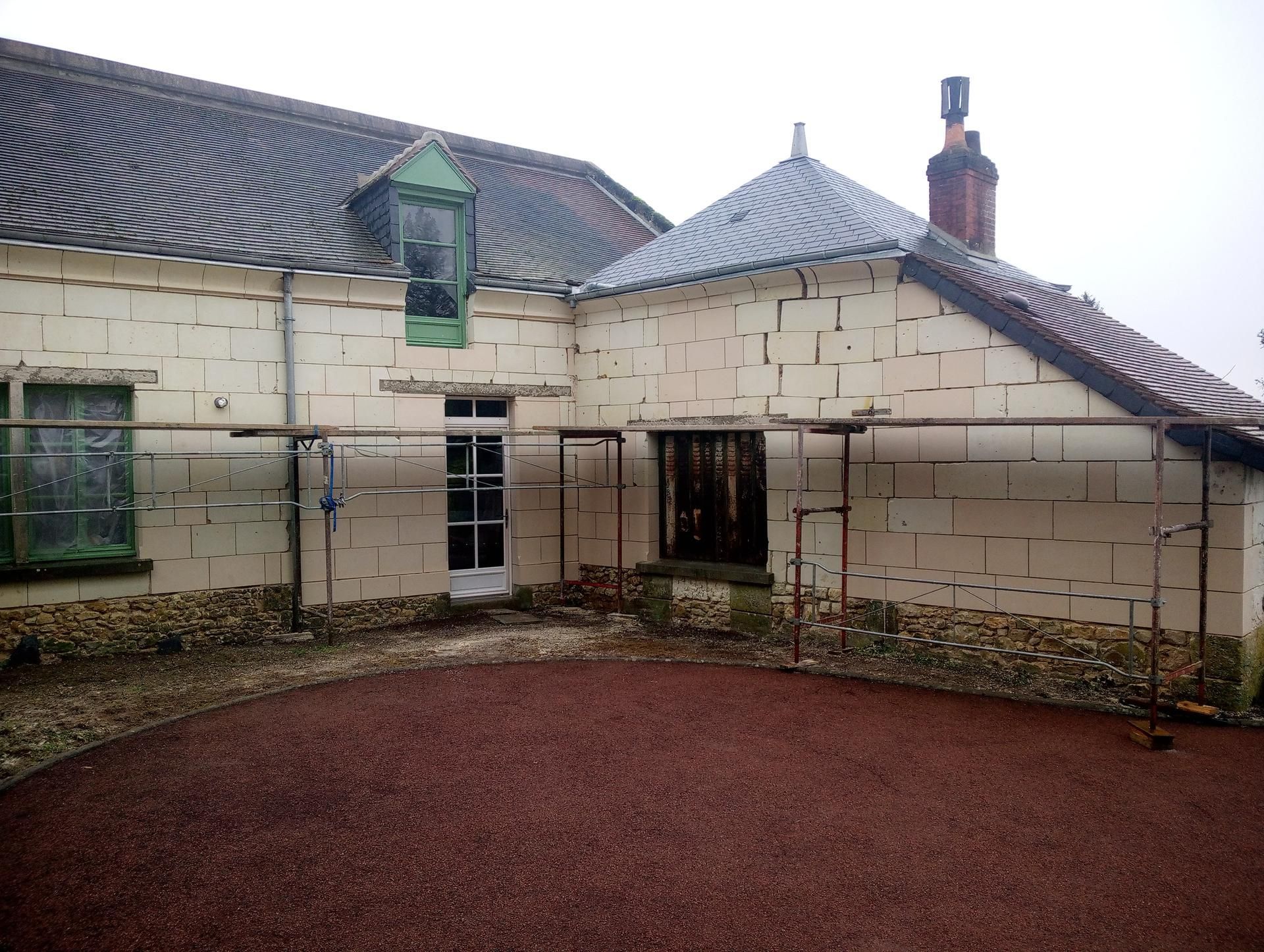 Bâtiment blanc au toit à motifs sombres, échafaudages et cour de gravier rouge.