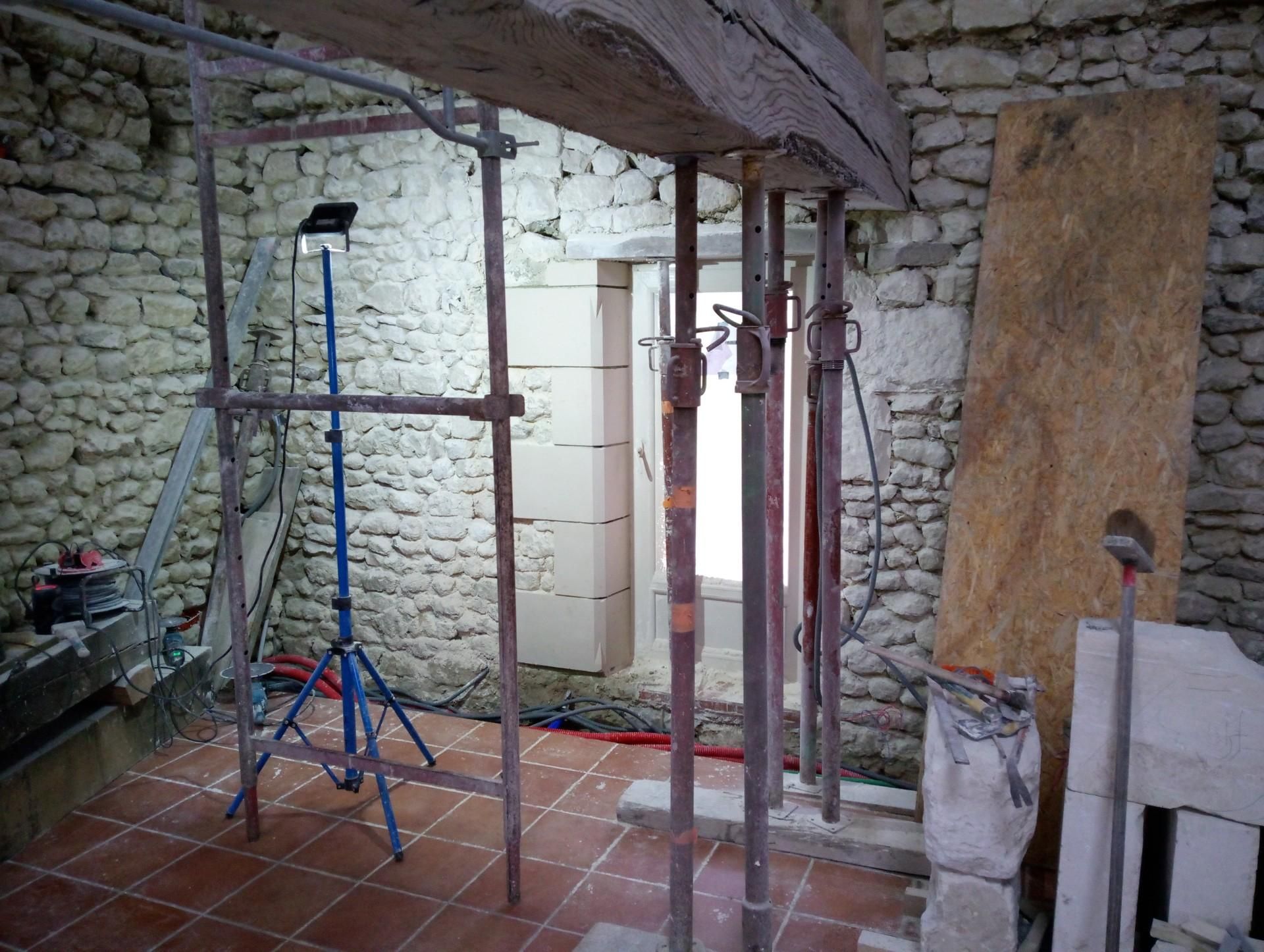Vue intérieure d'un chantier de construction. Poutre en bois soutenue par des vérins métalliques, murs de briques et de pierres visibles.