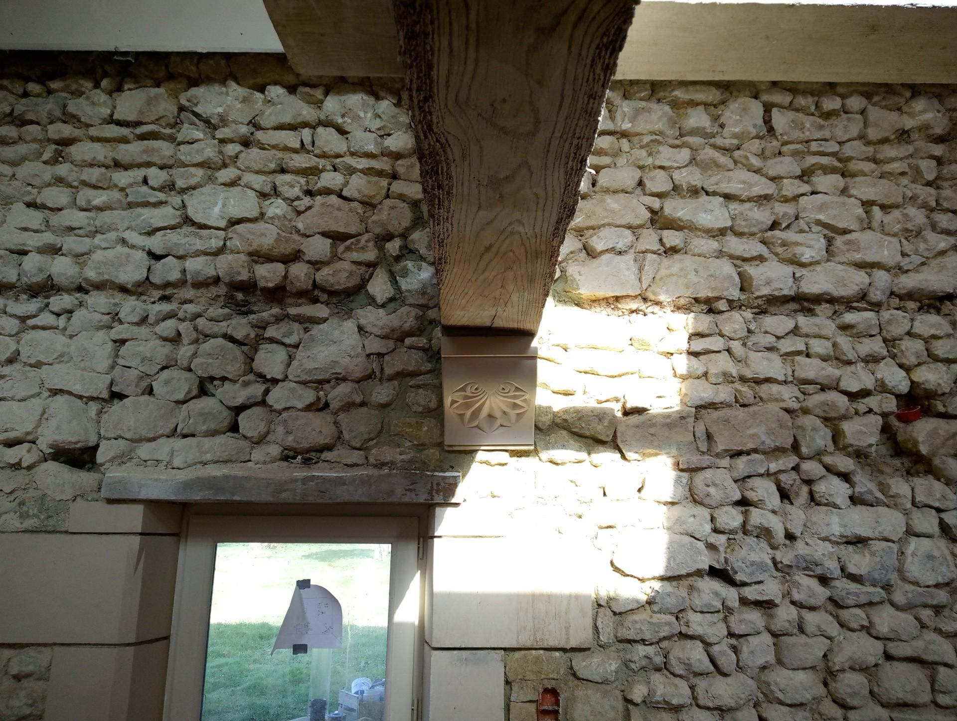 Mur en pierre avec poutre en bois et bloc décoratif au-dessus de la porte. La lumière du soleil projette des ombres.