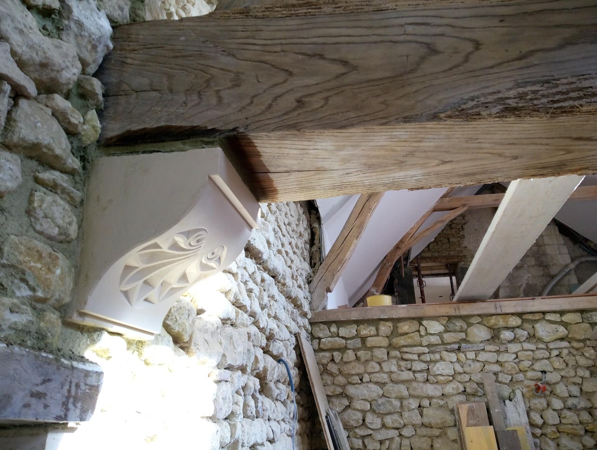 Mur en pierre avec poutre en bois, corbeau décoratif en plâtre et chevrons apparents.
