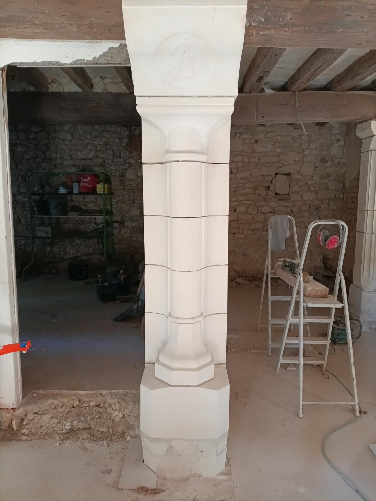 Colonne en calcaire en construction dans une pièce, soutenant des poutres en bois ; une porte et des outils sont visibles.