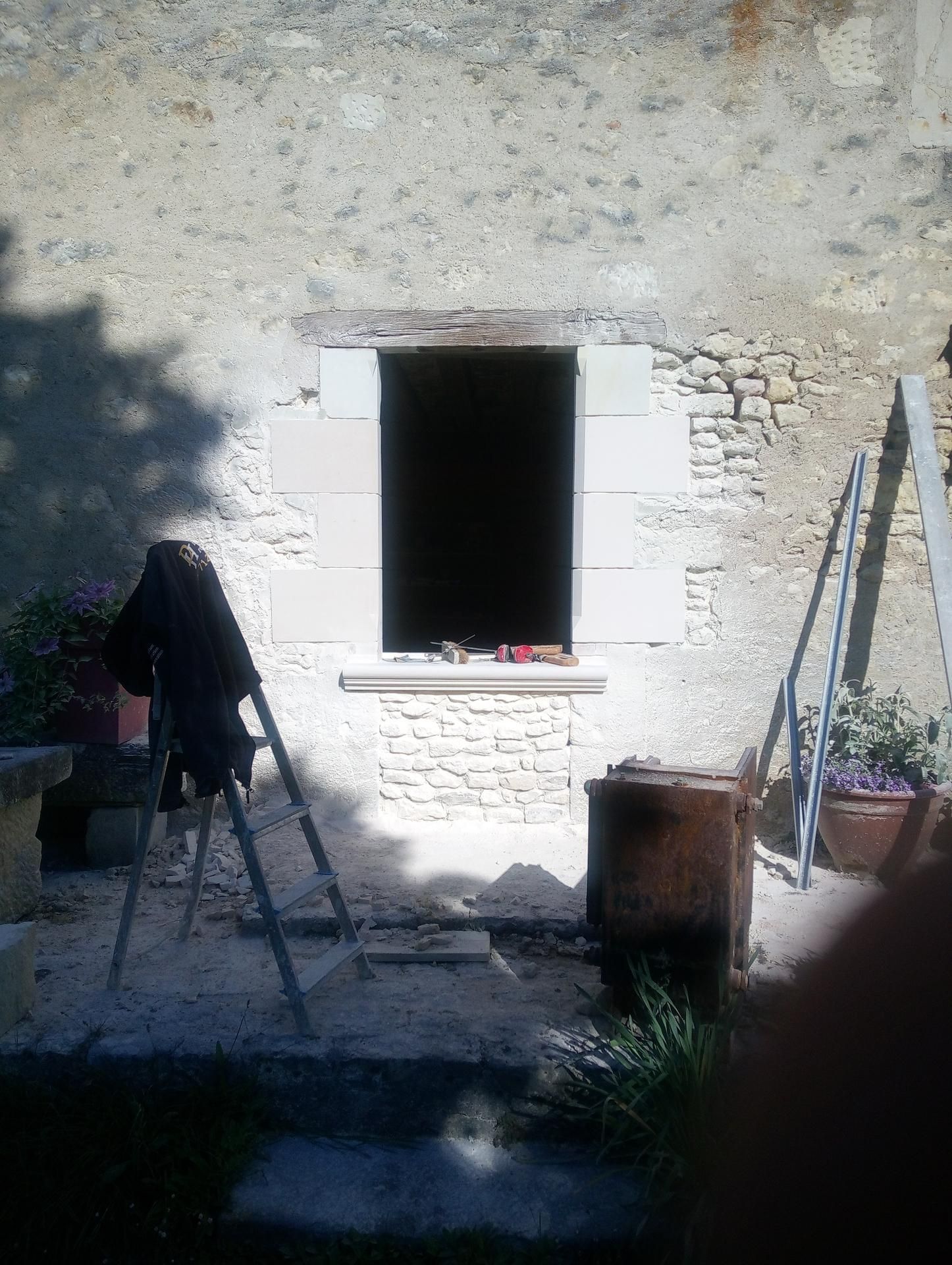 Fenêtre dans un mur de pierre, chevalet ombragé avec tissu, tonneau en bois et échelle à proximité.