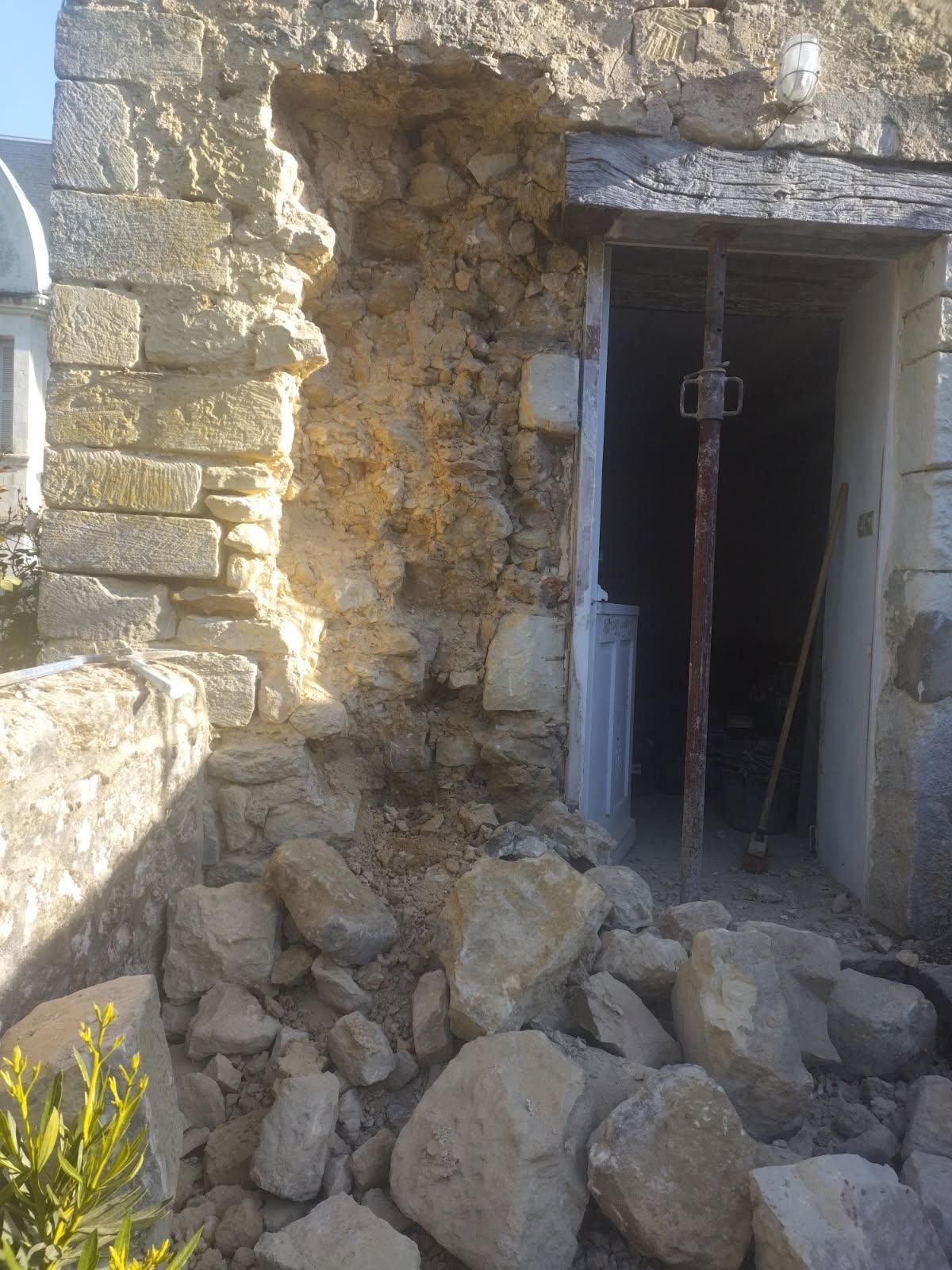 Mur en pierre endommagé avec un grand trou, gravats à la base, porte avec poutre de soutien visible.