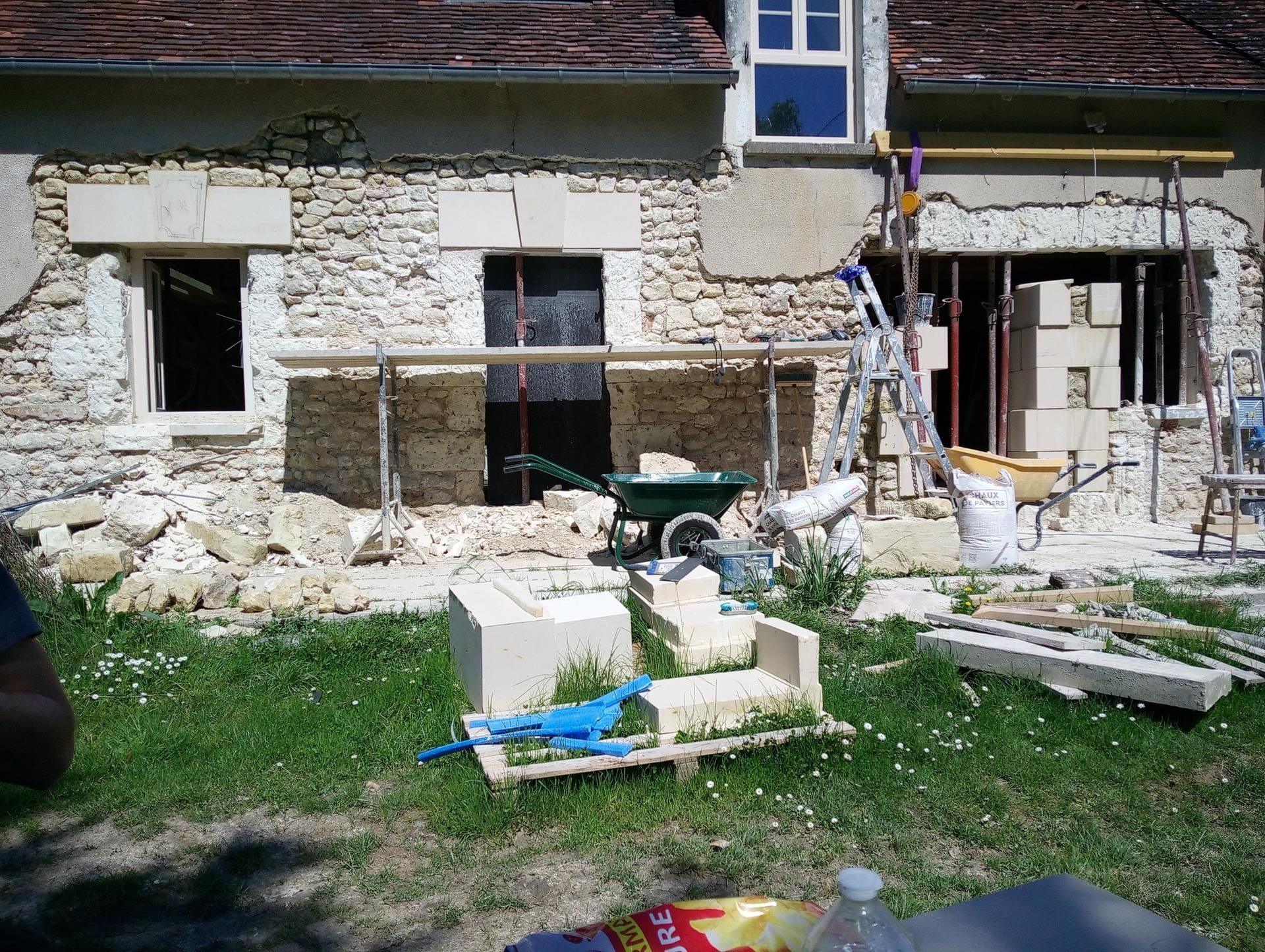 Maison en rénovation : façade en pierre, échafaudages, débris et outils dans une cour herbeuse.