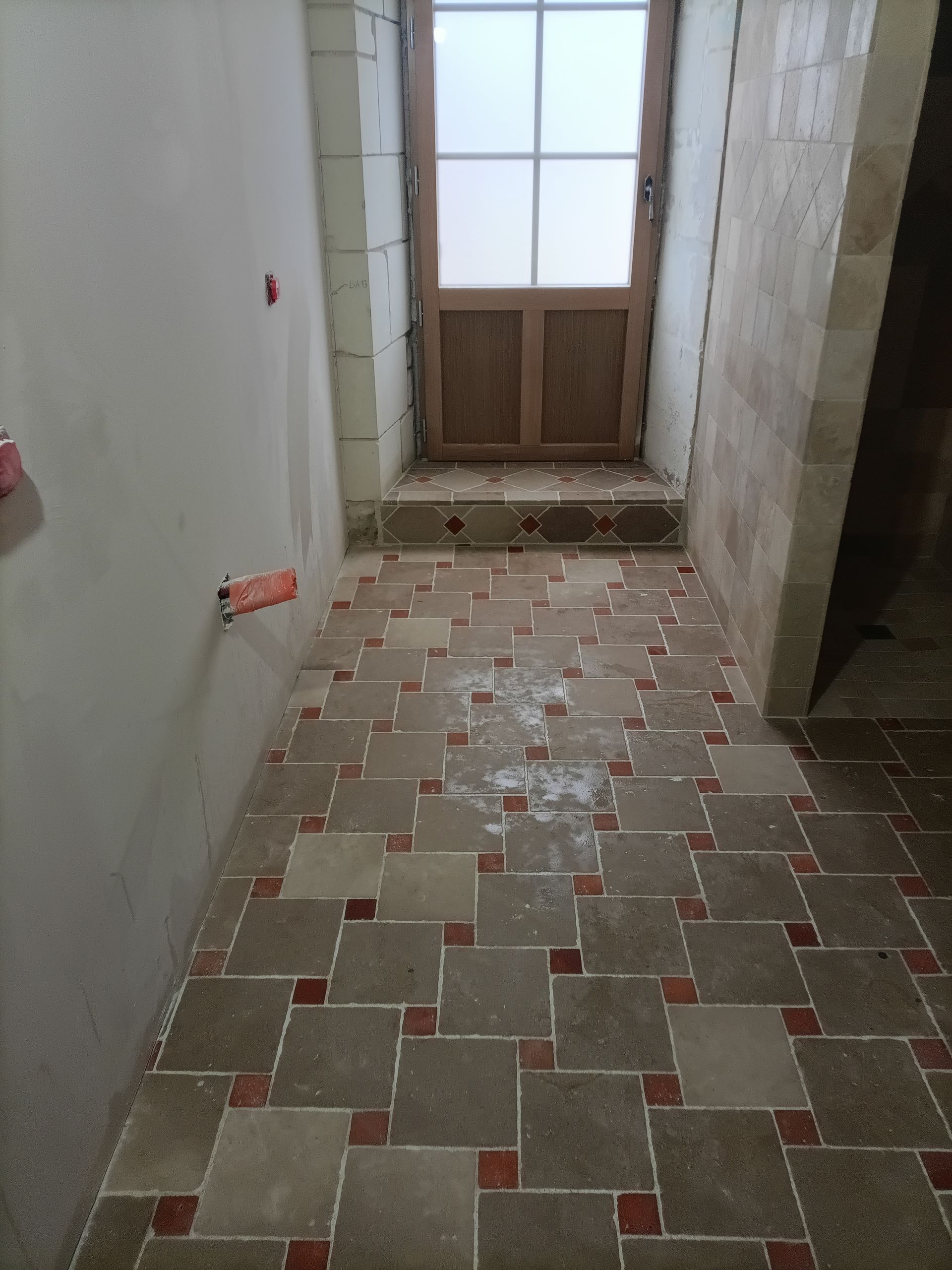 Couloir avec sol carrelé à motifs géométriques ; une porte en verre dépoli donne sur l'extérieur.