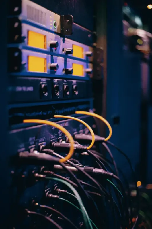 Un rack vertical con equipos de audio profesionales, pantallas iluminadas en amarillo y numerosos cables negros conectados.
