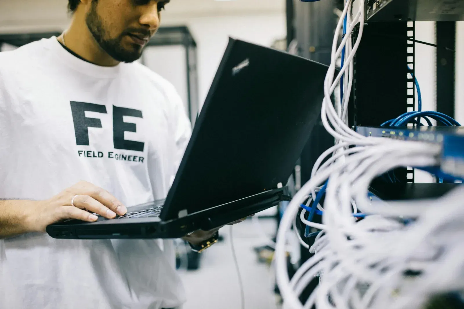 Un ingeniero de campo trabaja en un ordenador portátil frente a un rack de servidores de red con cables blancos y azules.