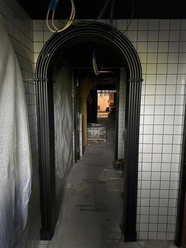 Schmaler Flur mit gewölbtem, schwarzem Türbogen. Weiss geflieste Wände. Schwach beleuchtet.