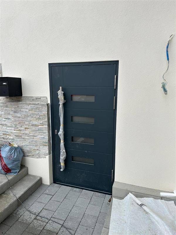 Graue Tür mit vier rechteckigen Glasscheiben, dekorativer Griff, an weisser Wand.