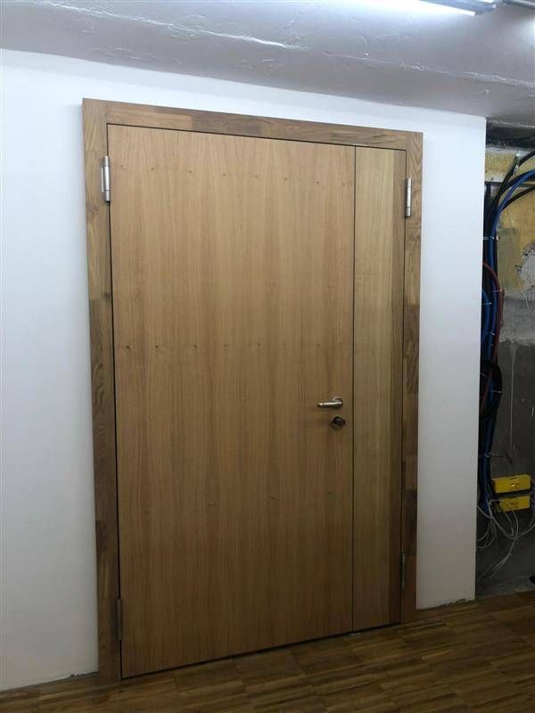 Eine Holztür in einem Holzrahmen vor weissen Wänden und einem Holzboden, mit sichtbaren Scharnieren und einem Türknauf.