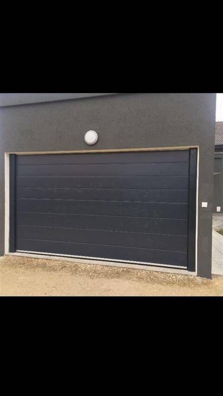 Dunkelgraues Garagentor an einem grauen Gebäude, darüber eine runde Leuchte.