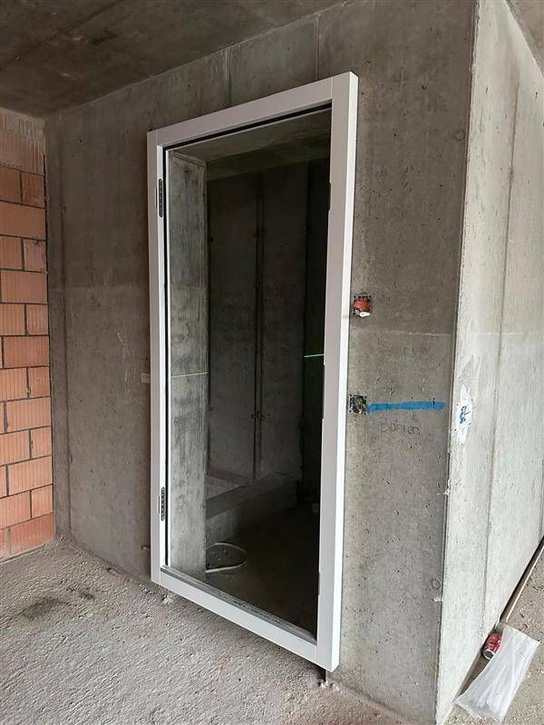 Tür mit weissem Rahmen in einer Betonwand, Rohbau. Blaue Leitung und roter Knopf sind an der Wand sichtbar.
